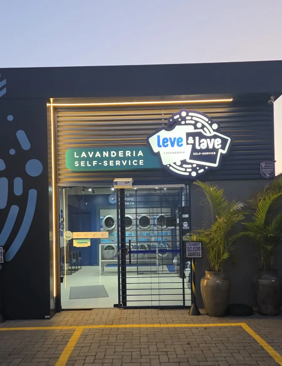 Fachada da Leve & Lave em Chapecó ao anoitecer: letreiro aceso, porta de vidro aberta e as máquinas visíveis da rua, com o estacionamento exclusivo na frente.