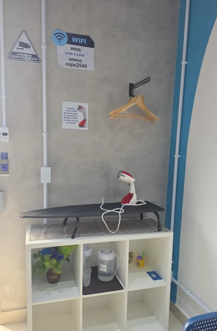 Bancada da Leve & Lave com tábua de passar, vaporizador e cabides de madeira liberados, ao lado do aviso com a rede e a senha do Wi-Fi.